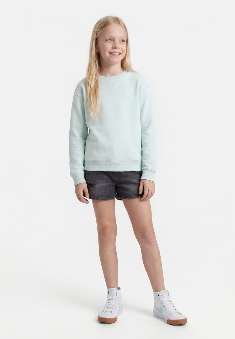 Νεαρή κοπέλα με μακριά ξανθά μαλλιά, που φοράει ανοιχτό μπλε φούτερ, μαύρο τζιν σορτς και λευκά ψηλά αθλητικά παπούτσια, χαμογελάει και στέκεται.