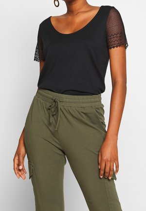 Vrouw draagt een zwarte top met een ronde hals en korte mouwen van kant, en olijfgroene broek met trekkoord en zakken aan de zijkant.