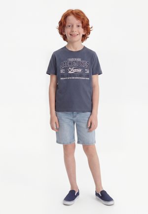 Rødhåret dreng smilende iført en blå grafisk t-shirt, lyse denimshorts og marineblå slip-on sneakers, stående mod en hvid baggrund.