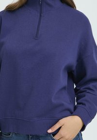 Vijoličast flis pulover z visokim ovratnikom in polnim zadrgo. Ima sproščen kroj in rebraste manšete, izdelan je iz mehkega, zračnega materiala.