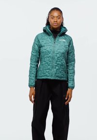 Vrouw staand, draagt een blauwgroene gewatteerde jas met capuchon en zwarte broek, tegen een effen lichte achtergrond.