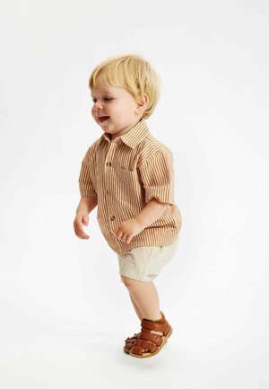 Camicia a maniche corte a righe in beige e ruggine, con taschino e chiusura a bottoni, abbinata a pantaloni corti beige chiaro e sandali marroni.
