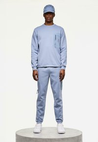 Hombre de pie en una plataforma redonda, vestido con un sudadera azul claro, joggers a juego con bolsillos con cremallera, gorra azul y zapatillas blancas.