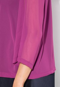 Lila Bluse aus einem halbtransparenten, leichten Stoff; mit langen, umgeschlagenen Ärmeln und einer leicht lockeren Passform. Glatte Textur und unaufdringliches Design.