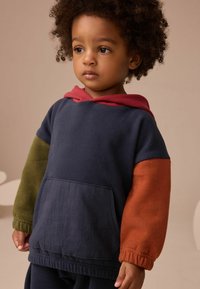 Maluch z kręconymi włosami, noszący granatowy hoodie z czerwoną kapturem, oliwkowym lewym rękawem i rdzawym prawym rękawem, patrzący nieco z boku.