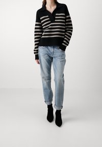 Suéter de punto a rayas en negro y beige con cuello estilo polo, combinado con jeans de mezclilla claros con dobladillo y botas negras de tobillo.