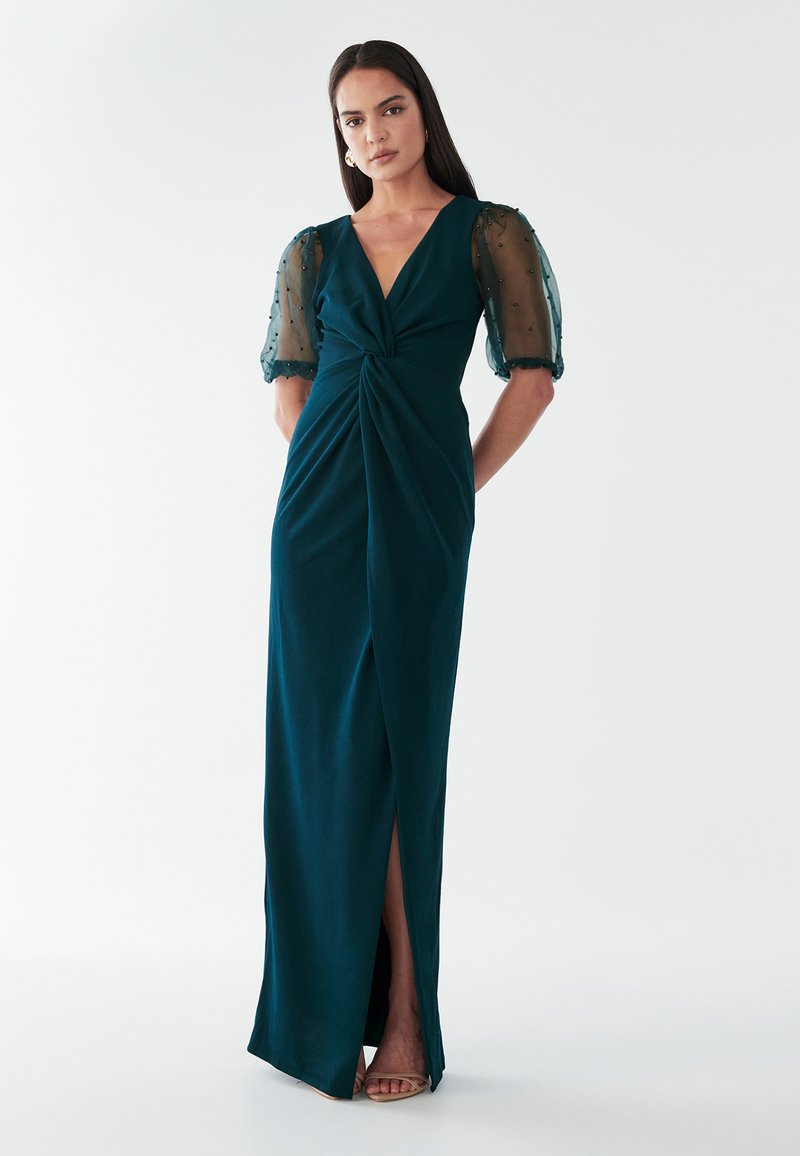 Teal jurk met een diepe V-hals, gedraaid voorkantdetail en doorzichtige puffmouwen. Heeft een hoge split tot de dij en een zachte, gladde stof.