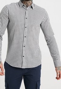 Geverifieerde button-up blouse in zwart-wit, lange mouwen, slim fit, met kraag en zwarte knopen tegen een ruitpatroon.