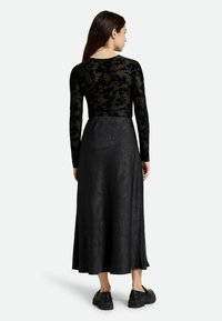 Schwarzes langärmliges Kleid mit einem durchsichtigen, gemusterten Oberteil und einem einfarbigen, strukturierten Rock. Der Rock hat ein dezentes Design und wird mit schwarzen Schuhen kombiniert.