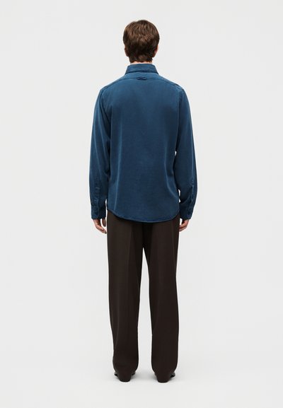 Άνδρας που στέκεται με πλάτη γυρισμένη, φοράει μπλε πουκάμισο με μακριά μανίκια και φαρδιά μαύρη παντελόνα, μπροστά σε απλό φωτεινό φόντο.