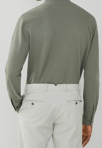 Chemise à manches longues de couleur olive avec une texture douce, associée à un pantalon gris clair doté de poches arrière et de boutons décoratifs.