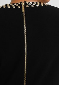 Schwarzes Kleidungsstück mit einem Reißverschluss auf der Rückseite, verziert mit metallischen goldenen Nieten am Ausschnitt für zusätzliche Details.