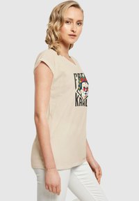 Beige bomulds t-shirt med korte ærmer, med et farverigt kranium grafik omgivet af teksten "Free Kahlo" i sort. Glat tekstur.