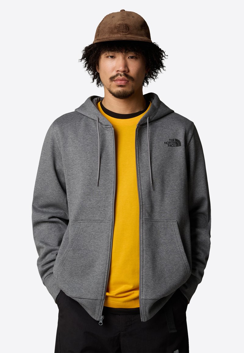 Sudadera con zip gris con dos bolsillos frontales, cordones y logo en el pecho, superpuesta a una camiseta amarilla con un cuello negro.