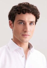 Camicetta bianca con colletto strutturato; presenta una fodera interna a motivi e bottoni perla. Tessuto liscio e design aderente.
