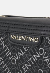 Borsa in tessuto nero con logo "Valentino" impresso in oro. Dettagli in pelle liscia sui bordi e chiusura con zip superiore. Sfondo con motivo geometrico.
