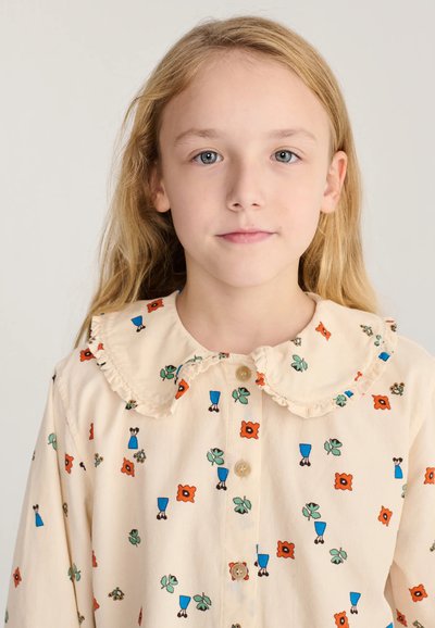 Jeune fille aux longs cheveux blonds portant un chemisier crème avec des motifs floraux colorés et un col à volants, regardant droit devant elle.
