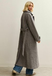 Abrigo largo de lana gris con cinturón, cuello y aberturas laterales. Presenta una tela texturizada y mangas amplias; combinado con unos vaqueros azules sueltos y zapatos ligeros.