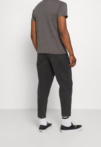 Persoon die een grijs T-shirt, zwarte jeans, witte sokken en zwarte sneakers draagt, staande tegen een effen witte achtergrond, met zijn rug naar de camera gekeerd.