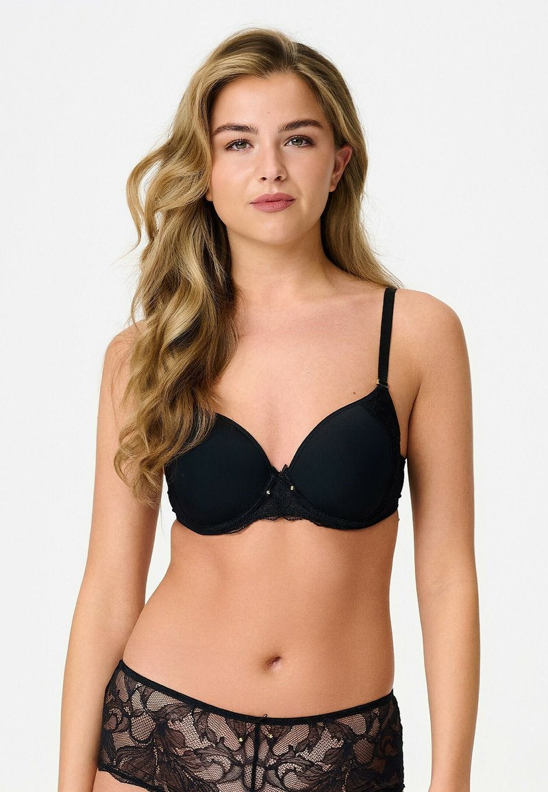 Jeune femme aux longs cheveux blonds ondulés portant un soutien-gorge noir rembourré et une lingerie en dentelle noire sur un fond blanc uni.