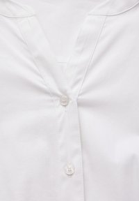 Weiße Baumwollbluse mit V-Ausschnitt, dezenten Falten und einer zentralen Knopfleiste mit einem runden, klaren Knopf. Glatte Textur.