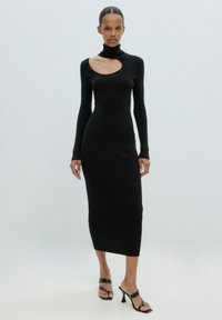 Schwarzes geripptes Kleid mit hohem Kragen und einem Schnitt auf der linken Schulter, taillierte Silhouette und knielange Länge. Getragen mit schwarzen hohen Sandalen.