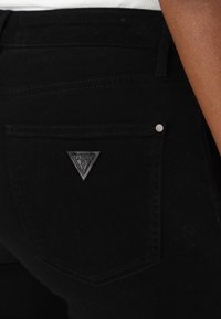 Melni džinsi ar trīsstūrveida Guess logo uz aizmugurējā kabatas, izturīga šūšana un gluda tekstūra.