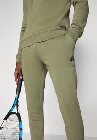 Olivengrønn hettegenser og joggebukser i myk, teksturert stoff; har svarte merkelapper, tettsittende design og sidelommer.