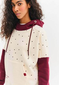 Bluza z kapturem w kolorze kremowym, z bordowymi rękawami, wzorem w kropki oraz przednią kieszenią. Posiada ściągacze i małą czerwoną metkę.