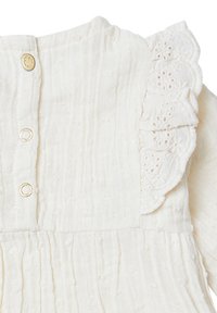 Vêtement blanc cassé en tissu texturé et froissé. Présente une bordure en dentelle festonnée et des fermetures à boutons dorés le long du dos.