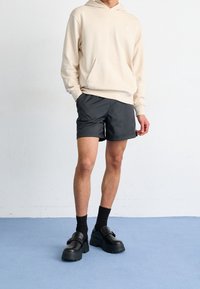 Felpa beige con cappuccio, pantaloncini neri, scarpe platform nere con fibbia e calzini neri. Design minimalista con vestibilità comoda e tessuto morbido.