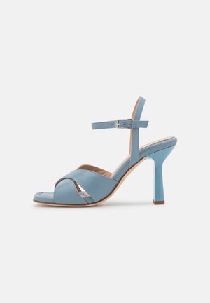Blå højhælede sandal med åbne tæer, krydsede frontstropper og en ankelrem med en sølvspænde på en hvid baggrund.