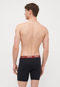 Sorte atletiske boxerbriefs med en rød Nike-logo på midjebåndet, der har en tætsiddende pasform og en glat tekstur. Designet er enkelt og minimalistisk.