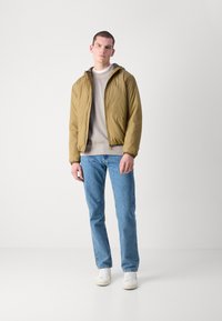 Khakifarbene Pufferjacke mit Kapuze, beiges Sweatshirt darunter, hellblaue Jeans und weiße Sneakers. Glatte Textur, lässiges Design.