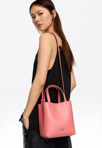 Bolso de mano de piel rosa con doble asa y una correa larga. Presenta una textura suave y un diseño minimalista, en contraste con un atuendo negro.