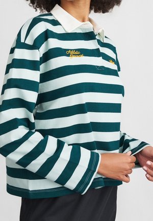 Osoba nosząca długą koszulę w turkusowo-białe paski z białym kołnierzem, żółtym haftowanym napisem i trzymająca mankiet koszuli.