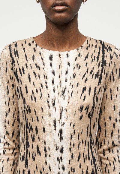 Γυναίκα που φοράει μπεζ μπλούζα με μαύρο και λευκό αφηρημένο μοτίβο με πινελιές και μικρούς χρυσούς κρίκους ως σκουλαρίκια.