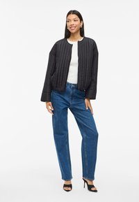 Juodas rištas bomberis su apvaliu iškirpimu ir ilgomis rankovėmis, derinamas su aukštu liemeniu mėlynomis tiesiomis džinsomis ir juodais aukštakulniais.