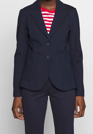 Persoon draagt een marineblauw colbert met twee knopen over een rood-wit gestreept shirt en bijpassende marineblauwe broek, staand tegen een effen achtergrond.