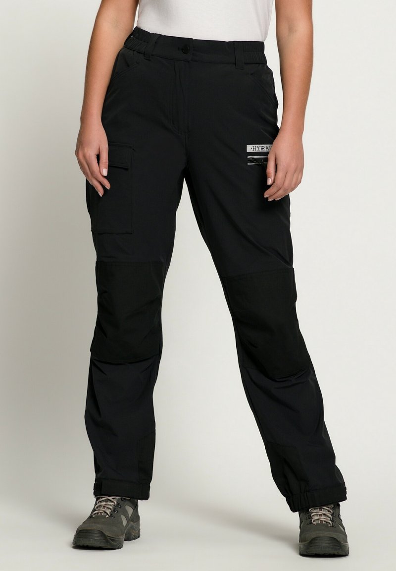 Femme portant un pantalon cargo noir avec des poignets élastiques et des chaussures de randonnée foncées, debout devant un fond clair uni.