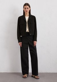 Zwarte cropped jacket met twee borstzakken, knoopsluiting en lange mouwen, gecombineerd met zwarte high-waisted wijde broek en sandalen.