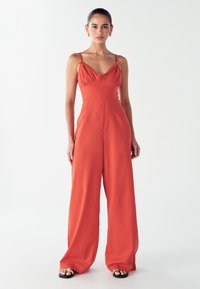 Žena v bezrukavnom širokom overal s rustikálno-oranžovou farbou, tenkými ramennými pruhmi a zlatými kruhovými náušnicami, stojí na bielom pozadí.