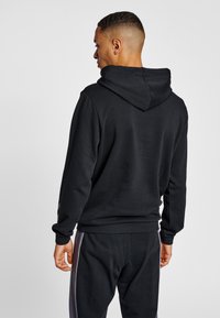 Svart hoodie med huva, tillverkad av texturerat tyg. Har ribbade muddar och fåll, med en logodetalj på vänster ärm.