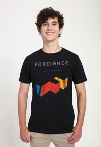 Černé bavlněné tričko s nápisem "FOREIGNER" nad geometrickým vzorem v modrých, červených a žlutých tvarech. Krátké rukávy a kulatý výstřih.