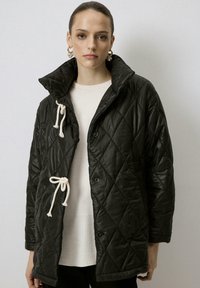 Chaqueta negra acolchada con un alto cuello, que presenta cordones y botones color crema. Textura suave, tejido ligero y ajuste holgado.