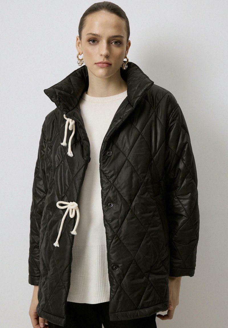 Chaqueta negra acolchada con un alto cuello, que presenta cordones y botones color crema. Textura suave, tejido ligero y ajuste holgado.