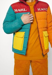 Cazadora acolchada colorida con paneles en rojo, verde azulado y naranja. Presenta bolsillos de parche amarillos y la palabra «KARL» bordada en el pecho. Tejido texturizado.