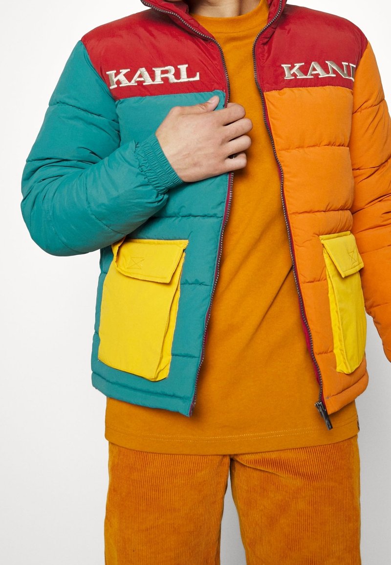 Cazadora acolchada colorida con paneles en rojo, verde azulado y naranja. Presenta bolsillos de parche amarillos y la palabra «KARL» bordada en el pecho. Tejido texturizado.