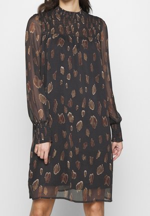 Kvinde iført en sort kjole med brune abstrakte pletter, lange transparente ærmer samt smockede manchetter og udskæring.