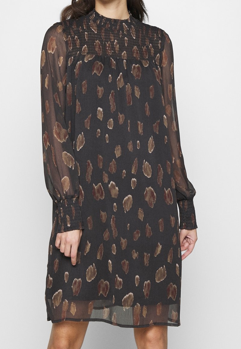 Femme portant une robe noire avec des taches abstraites marron, des manches longues transparentes, et des poignets et un col froncés.